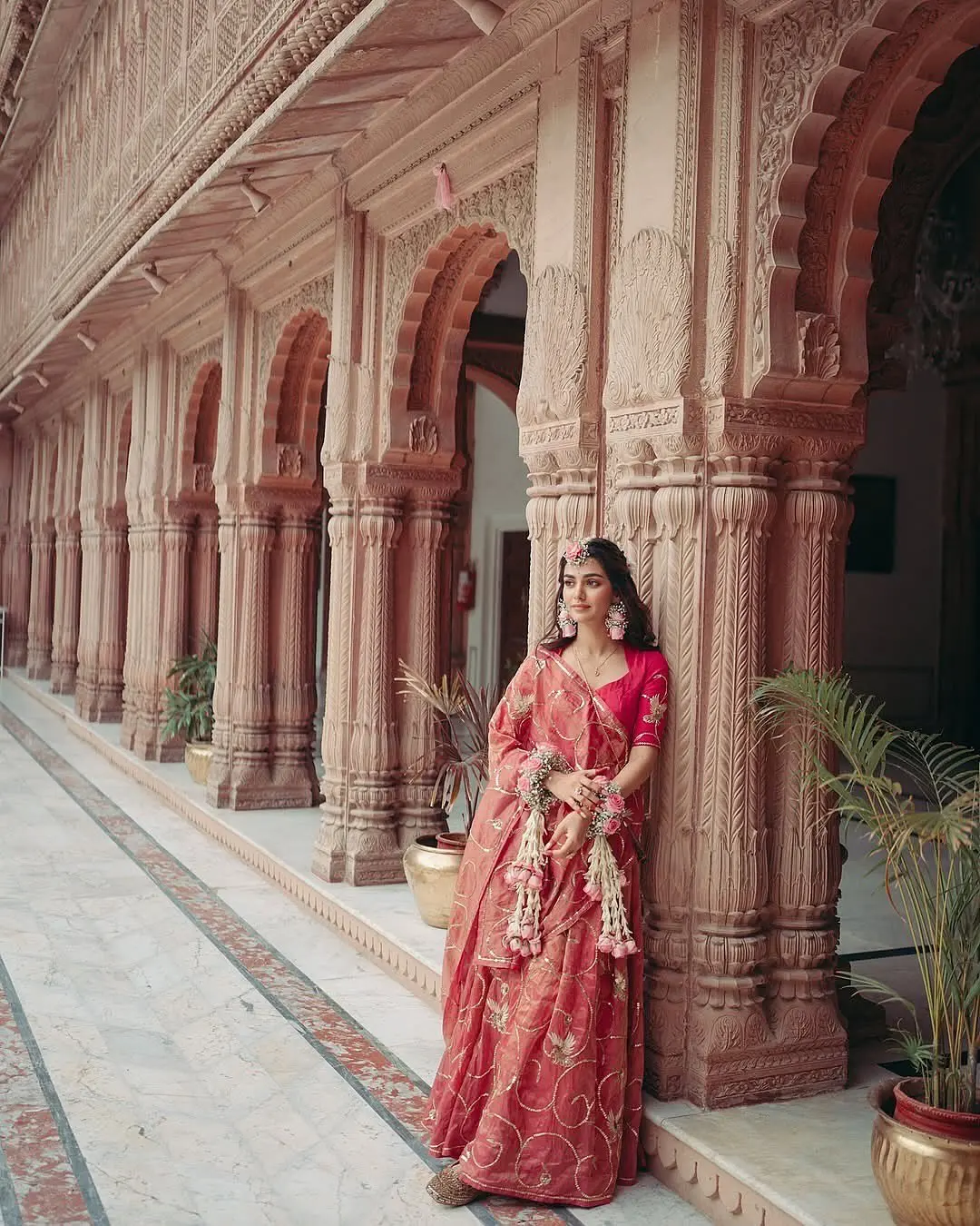 Editorial-style bridal portrait of a bride in a deep pink lehenga leaning against an intricately carved heritage palace pillar.