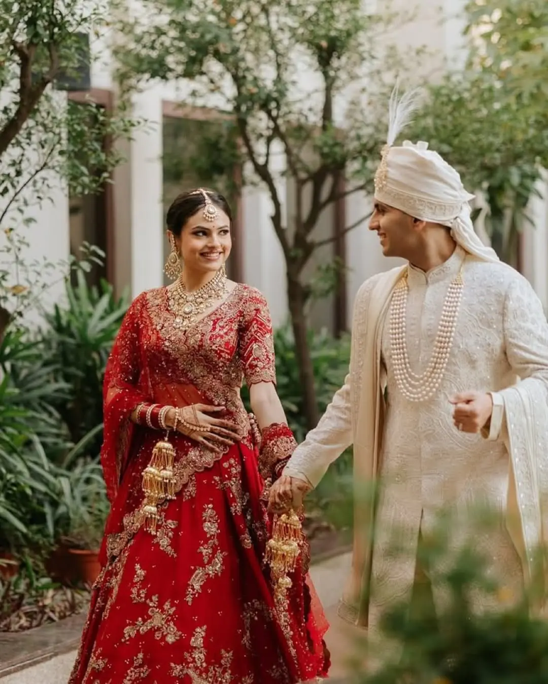 Authentic candid wedding photography of a happy Indian couple walking together outdoors, featuring a red bridal lehenga and ivory groom's sherwani.