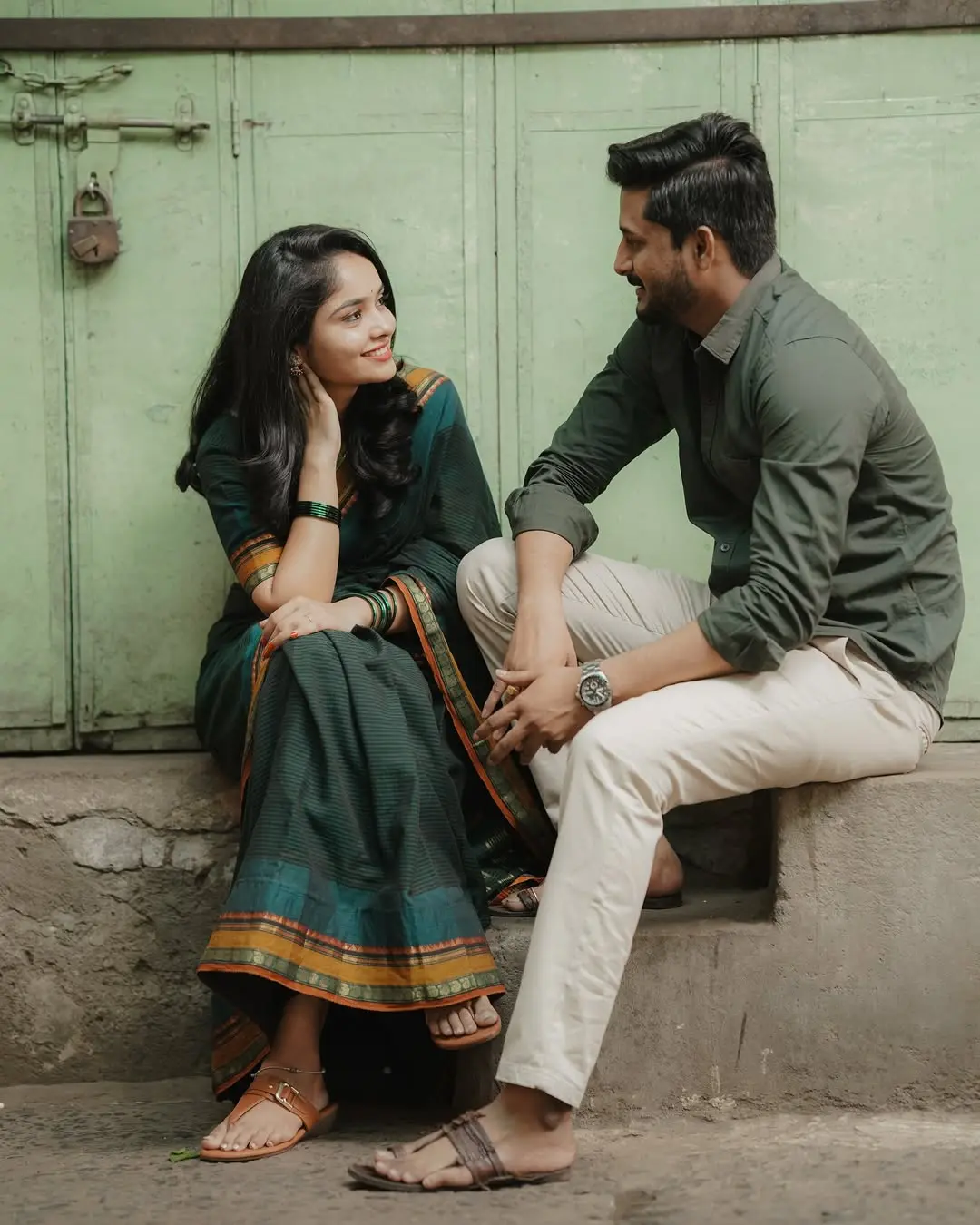 Editorial-style pre-wedding portrait of a couple in traditional Indian attire sitting against a rustic green door, capturing quiet luxury.